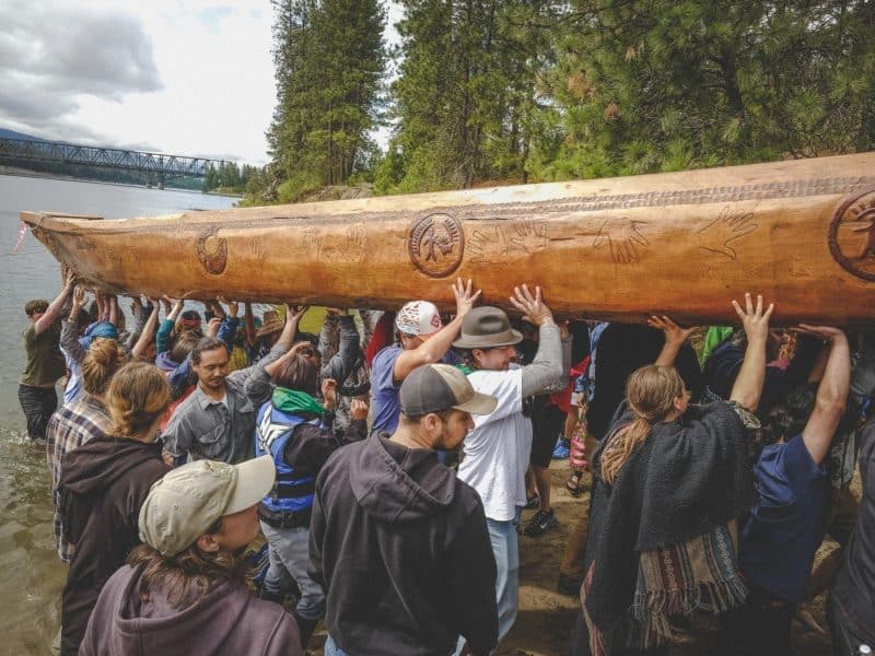 More than a dozen people are carrying a large carved wooden canoe from the water.