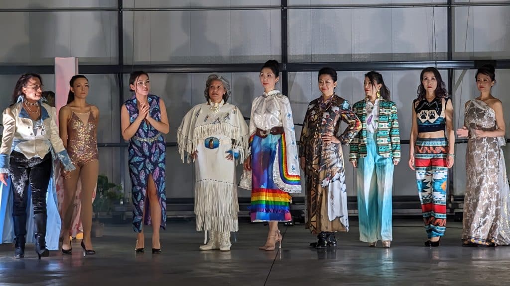 The photo shows the fashion designer to the far right and a line up of models dressed in her clothing. The collection ranges from more modern clothing to traditional buckskin regalia. The clothing is speckled with vibrant and bright patterns. 