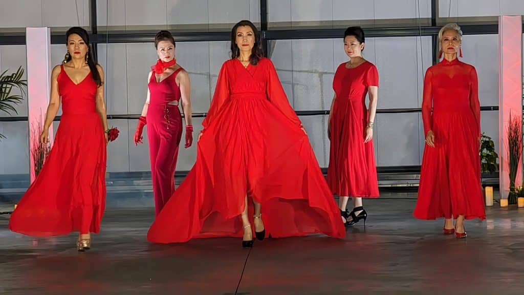 The photo shows five models wearing stunning and vivid red dresses. Two of the models are standing further back, and three of the models are walking towards the camera. 