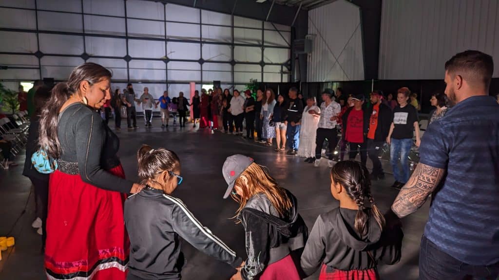 The photo shows dozens of people joined in hands for a round dance. In the foreground, there are three young children, with a woman to their left and a man to their right. They are wearing red under their jackets. 