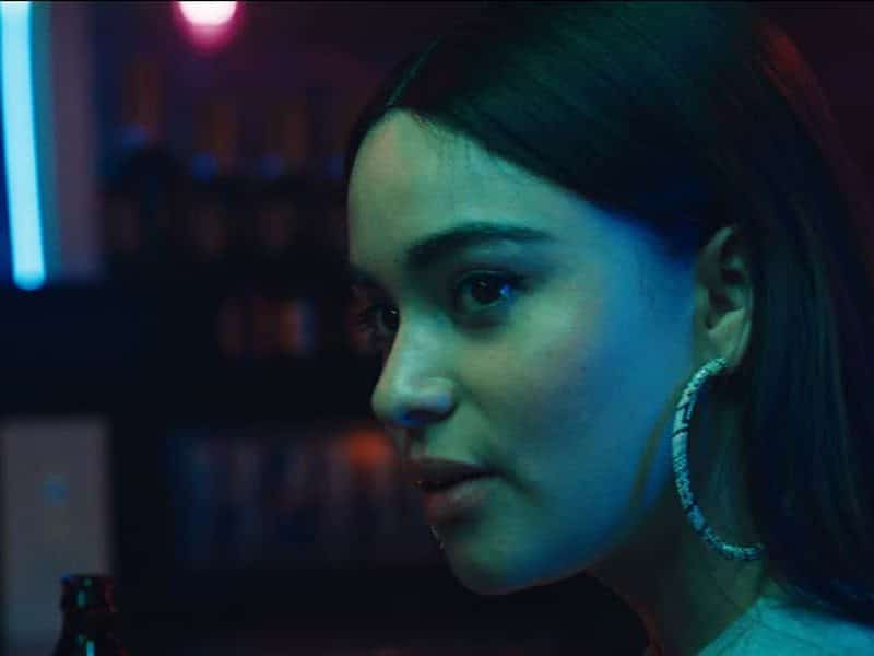 Photo shoes a woman lit by a blue-green neon light. Her face is in frame and the background is fuzzy and dark.