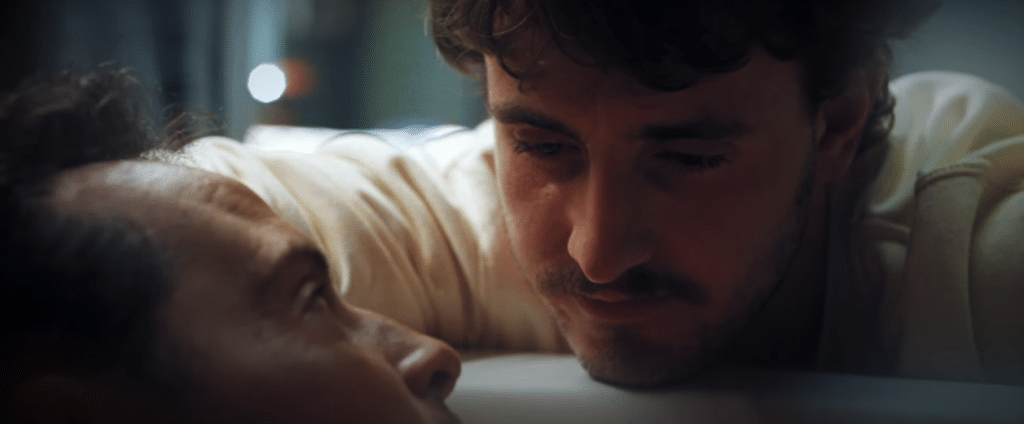 Photo shows two men, with one man lying down and his face cut off by the bottom of the photo. The second man is looking down tenderly at him and lit in a soft light.