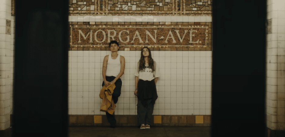Two people are in the center of the photo leaning back against a subway tiled wall. Above their heads, the tile is inlaid with the location, reading MORGAN-AVE. There are two dark pillars on each side of them, presumably from across the subway, where the camera is positioned.