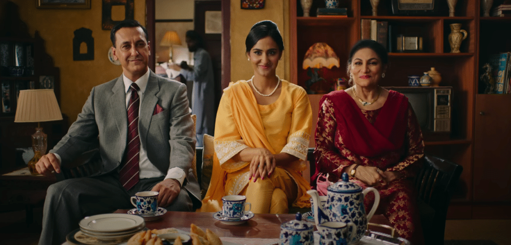 A man and two women are seated on a couch, with a beautiful antique tea set on the table in front of them. They are are smiling, but in a polite and professional way.