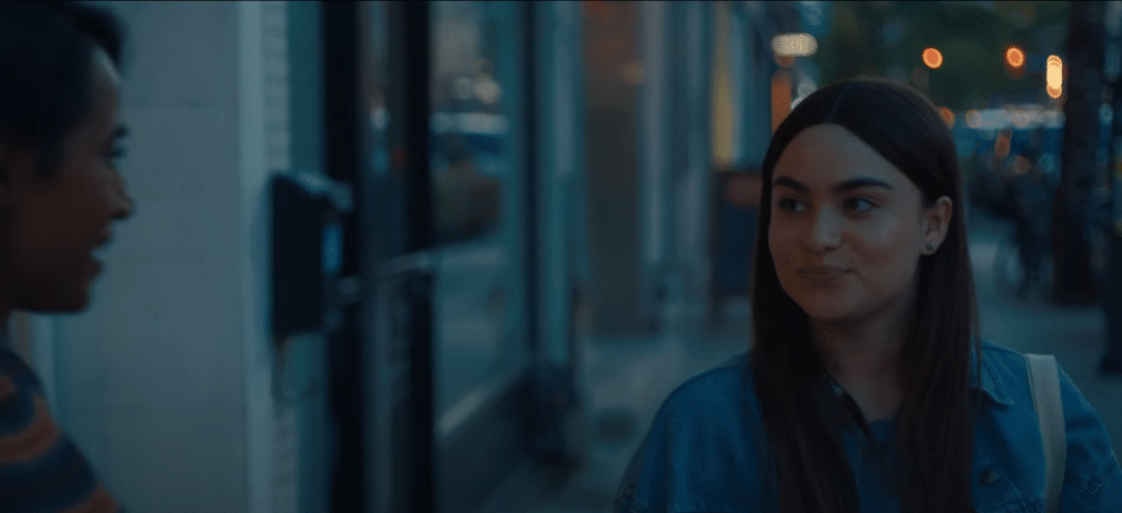 Two women are smiling at each other. The first woman is facing away from the camera, and is slightly cut off from the left side of the photo. The other woman is facing towards the camera. They are on a street sidewalk at dusk.
