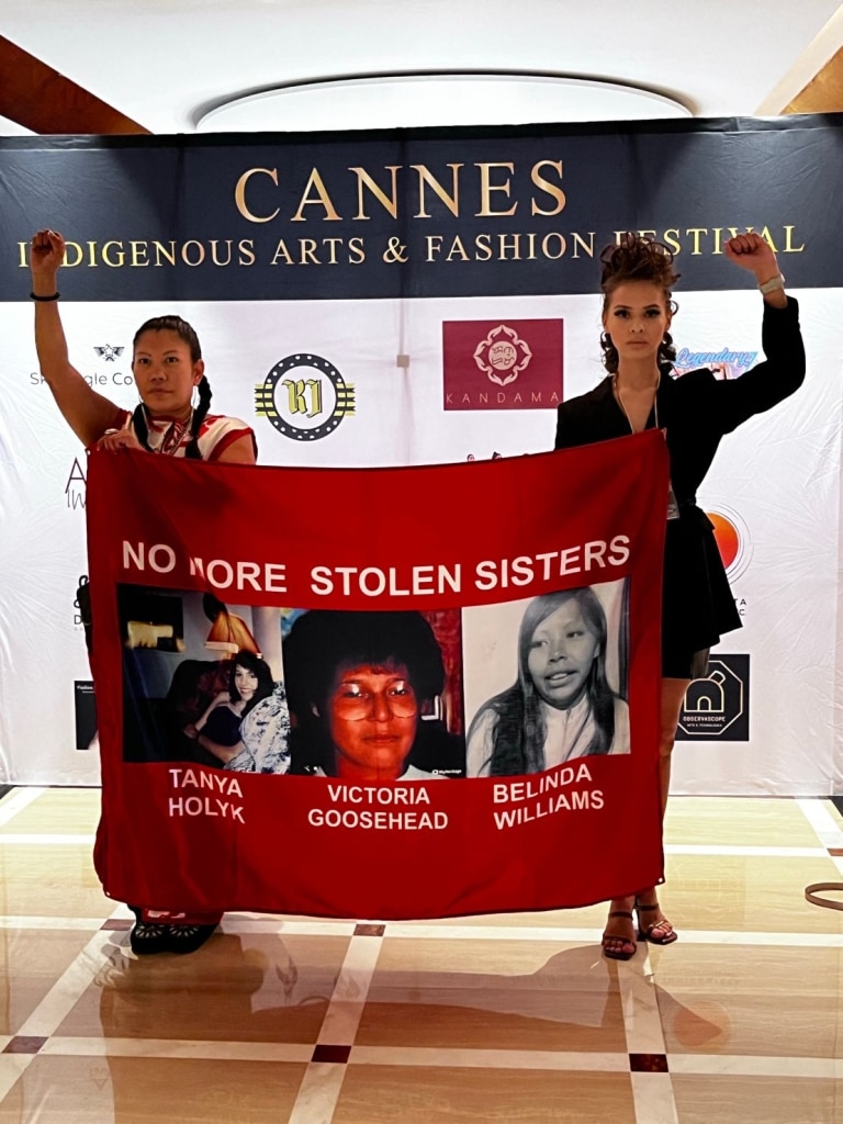 Two women are standing with their fist raised in a clench. They are holding a banner that says "No More Stolen Sisters" with photos of three women on it. Their names are beneath each photo and read "Tanya Holyk", "Victoria Goosehead", and "Belinda Williams" respectfully. On the backdrop behind the women is a banner that reads "Cannes Indigenous Arts and Fashion Festival"