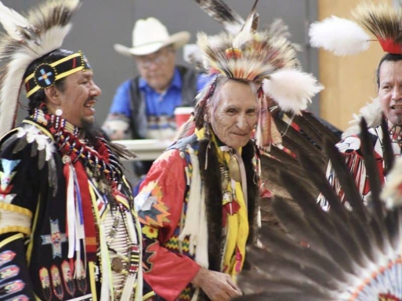 Three men are standing in the photo, all dressed in men's traditional Indigenous regalia.