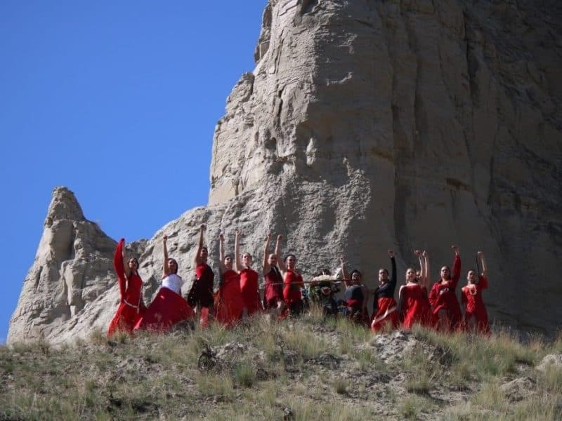 A line of women dressed in red all have one hand raised with thier fist clenched. In front of them, there is a man in traditional Indigenous regalia seated lower in the grass. The background is the iconic hoodoos located between the Tk'emlups reserve and the LaFarge bridge.