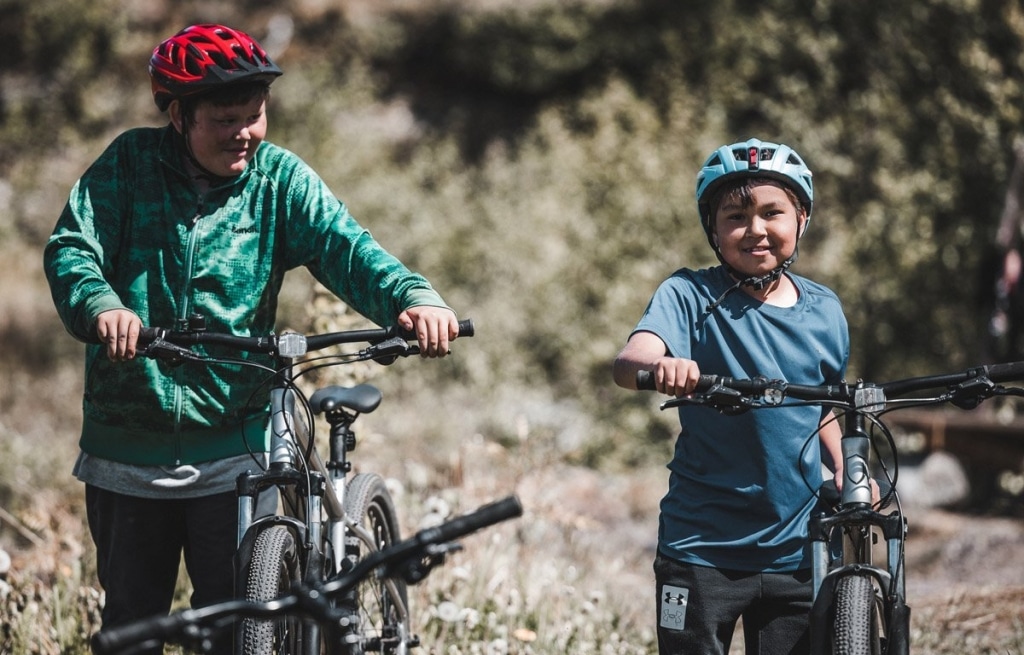 Photo shows two young children, possibly around the ages of ten to fourteen, pushing their mountain bikes and smiling.