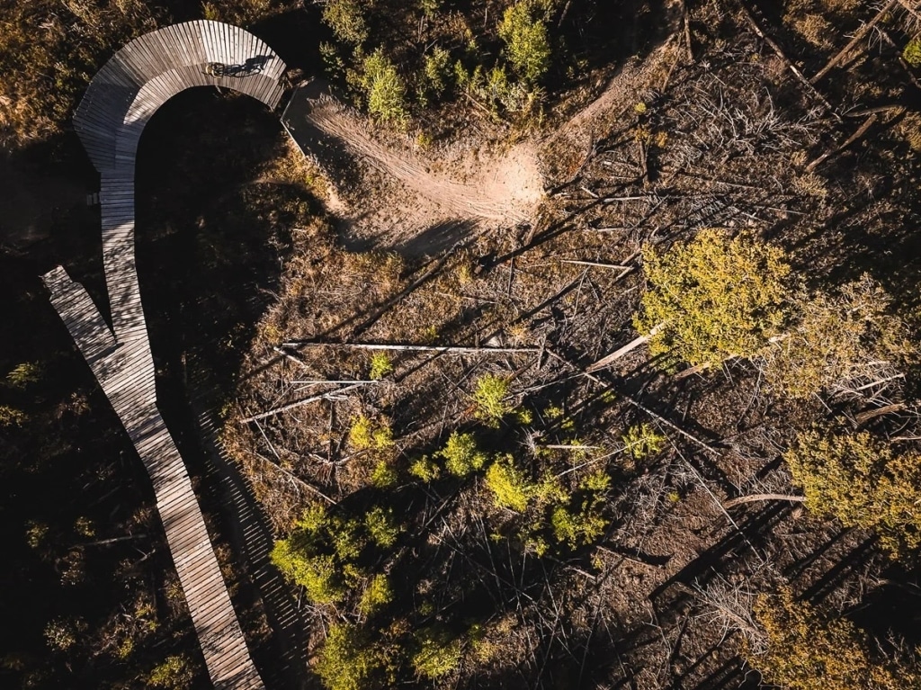 Arial shot shows a rugged mountain biking path from a bird's eye view. There is a wooden ramp to the left side of the photo.