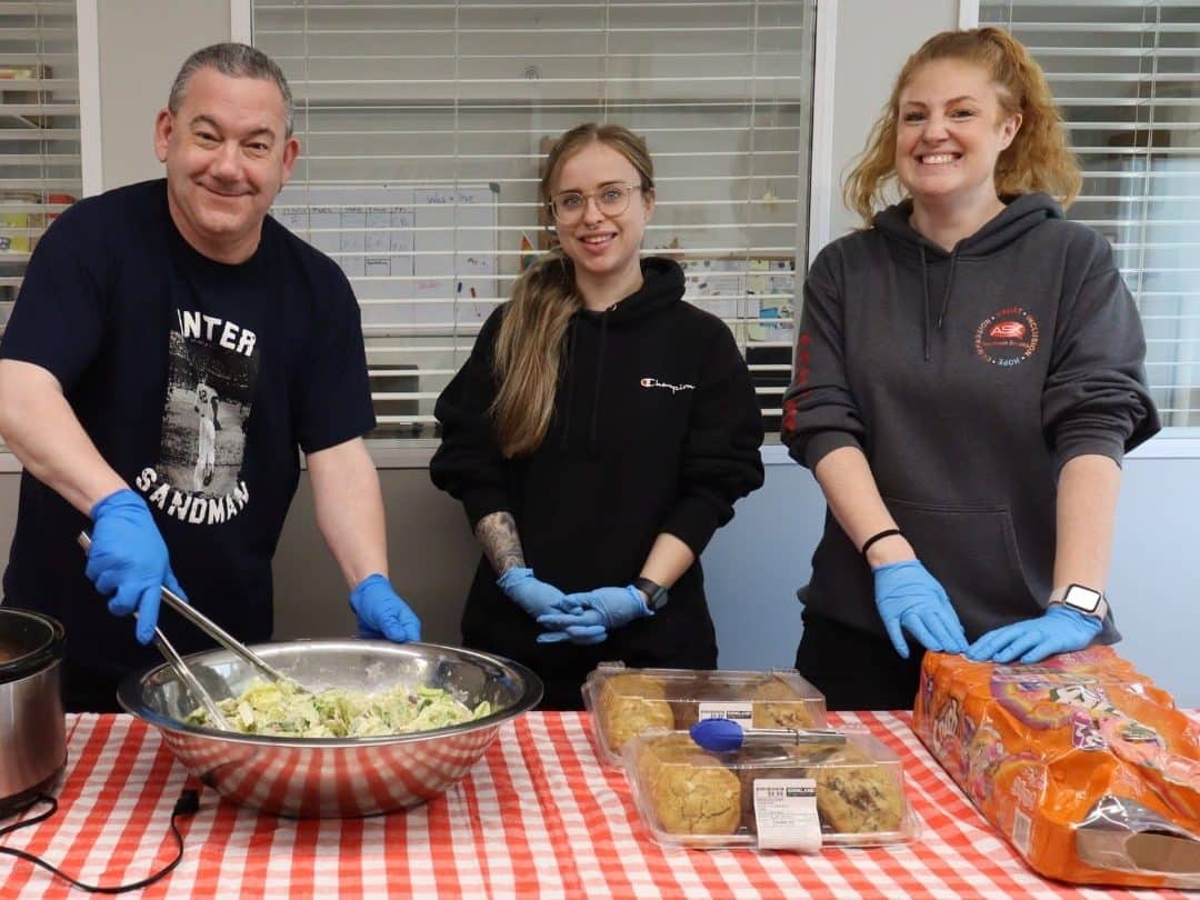 Three people with gloves on prepare food for a community meal.