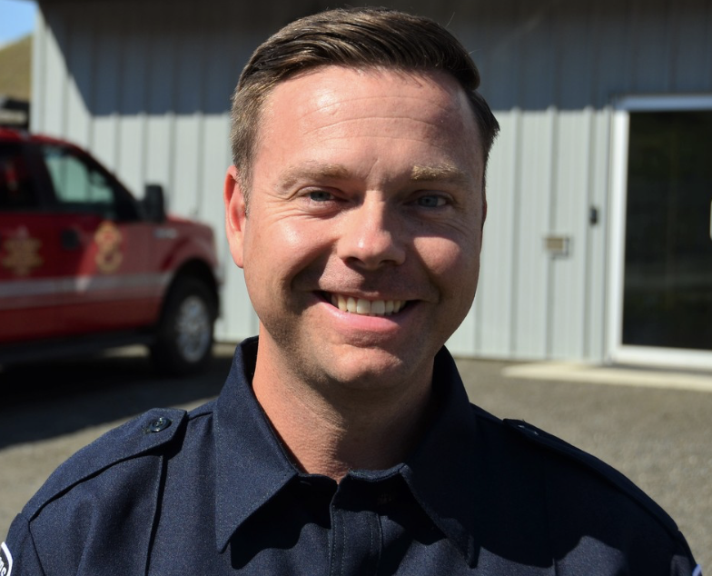A man in fire rescue uniform stands in front of the station sunlit.