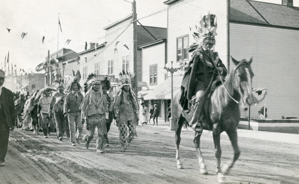 Kamloops historical parade photos show a chief in headdress riding a horse along an old street with a procession of Secwepemc in traditional dress walking behind
