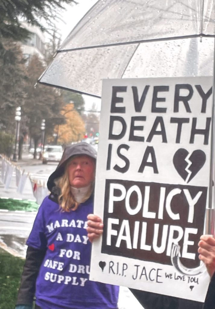 A woman stands in the rain holding a poster that reads "Every death is a policy failure"