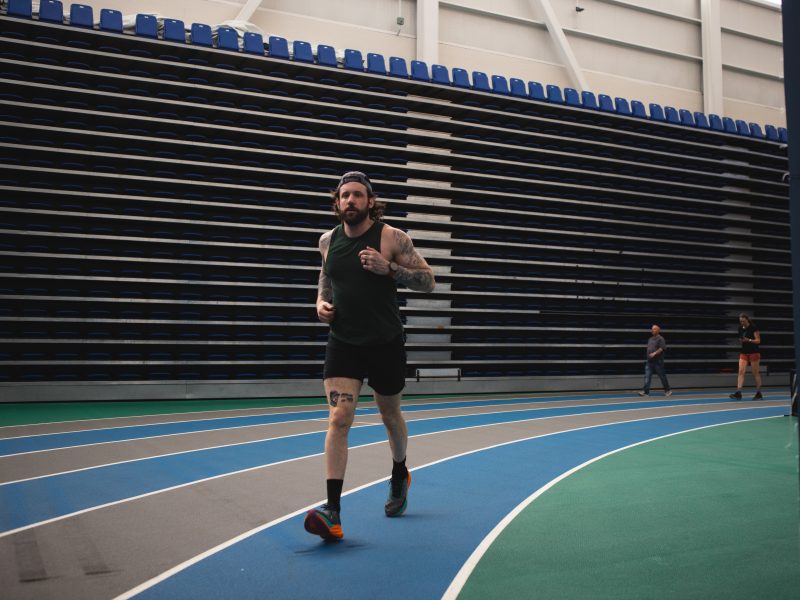 A man is seen running in the indoor track, the track has 6 paths each of them has alternating colors of grey and blue. The man is running on a blue track