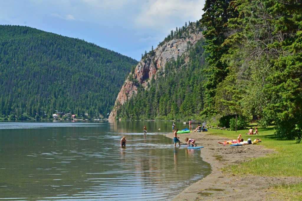 Photo of Paul Lake surrounded by trees. There is a beach where people are in the water and enjoying the sun. Camping sites near Kamloops